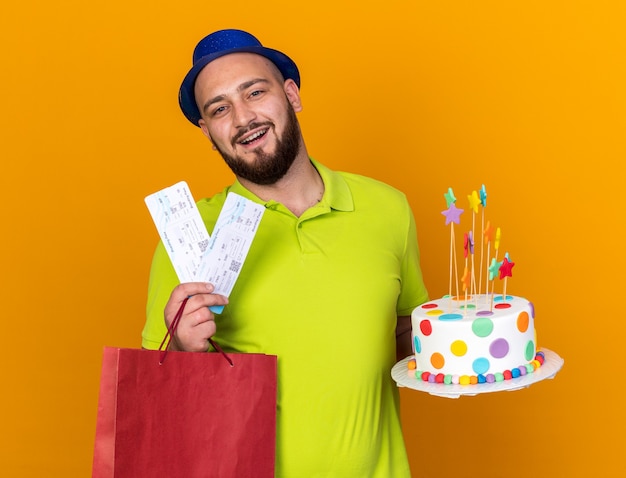 Sorridente giovane uomo che indossa cappello da festa tenendo la borsa regalo con torta e biglietti isolati su parete arancione