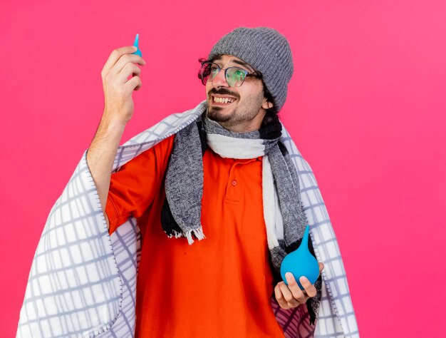 Sorridente giovane uomo malato con gli occhiali inverno cappello e sciarpa avvolti in plaid tenendo i clisteri guardando il piccolo isolato sulla parete rosa con lo spazio della copia