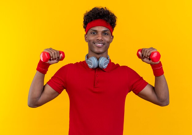 Sorridente giovane uomo sportivo afro-americano che indossa la fascia e il braccialetto in cuffie sul collo che alza i manubri isolati sulla parete gialla