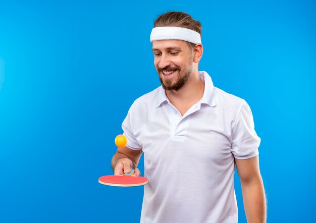 Sorridente giovane uomo sportivo bello che indossa la fascia e braccialetti lanciare la palla con la racchetta da ping pong e guardarlo isolato su spazio blu