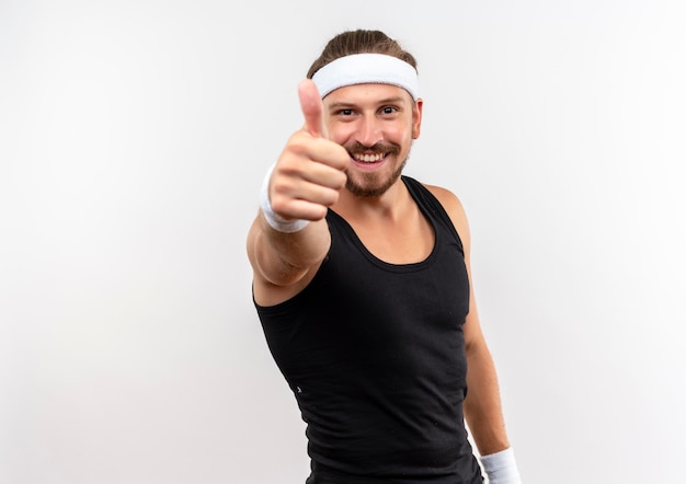 Sorridente giovane uomo sportivo bello indossando la fascia e braccialetti che mostrano il pollice in su isolato su spazio bianco