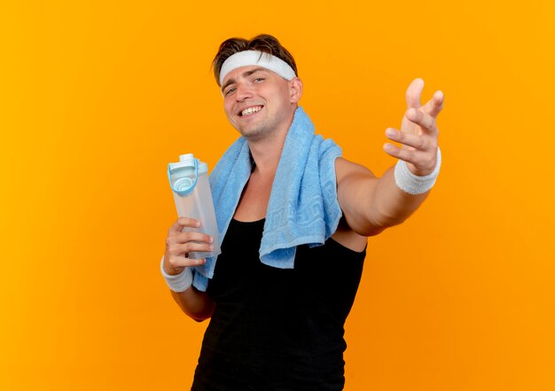 Sorridente giovane uomo sportivo bello indossando la fascia e braccialetti che tengono la bottiglia di acqua isolata sulla parete arancione
