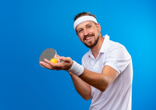 Sorridente giovane uomo sportivo bello indossando la fascia e braccialetti in possesso di racchette da ping pong con palla isolato su spazio blu