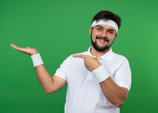 Sorridente giovane uomo sportivo che indossa la fascia e il braccialetto che finge di tenere e indica qualcosa