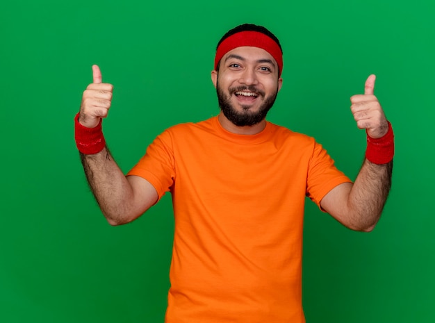 Sorridente giovane uomo sportivo che indossa la fascia e il braccialetto che mostra i pollici in su isolato su sfondo verde