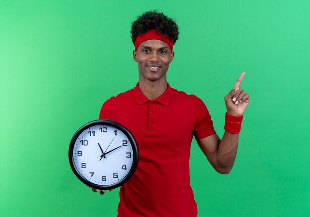 Sorridente giovane uomo sportivo che indossa la fascia e il braccialetto che tiene orologio da parete