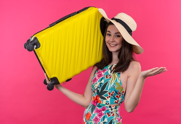 Sorridente giovane viaggiatore donna che indossa il cappello che tiene la valigia sulla parete rosa isolata