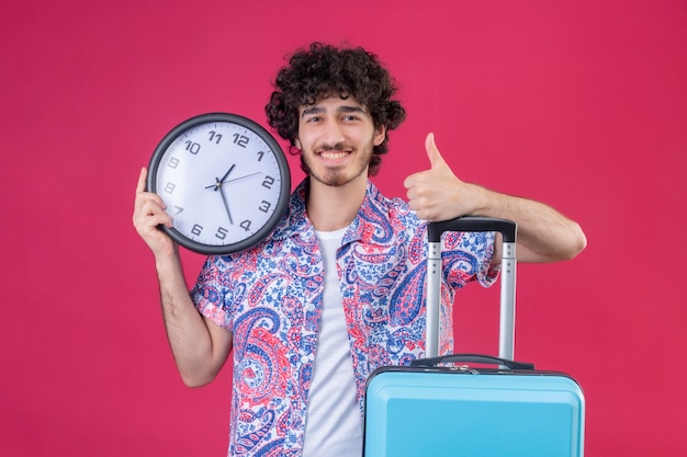 Sorridente giovane viaggiatore riccio bello uomo che tiene l'orologio che mostra pollice in alto mettendo la mano sulla valigia sulla parete rosa isolata