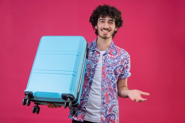 Sorridente giovane viaggiatore riccio bello uomo che tiene la valigia che mostra la mano vuota sulla parete rosa isolata
