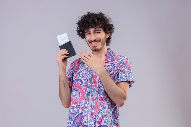 Sorridente giovane viaggiatore riccio bello uomo che tiene portafoglio e biglietti aerei e indicando con la mano su di esso sulla parete bianca isolata con lo spazio della copia
