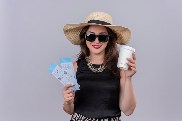 Sorridente giovane viaggiatrice che indossa canottiera nera in cappello e bicchieri in possesso di biglietti e una tazza di caffè sul muro bianco