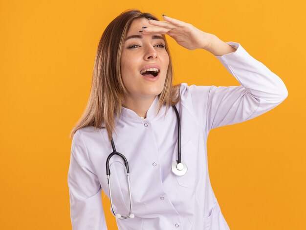 Sorridente guardando al lato giovane medico femminile che indossa abito medico con lo stetoscopio guardando a distanza con la mano isolata sulla parete gialla