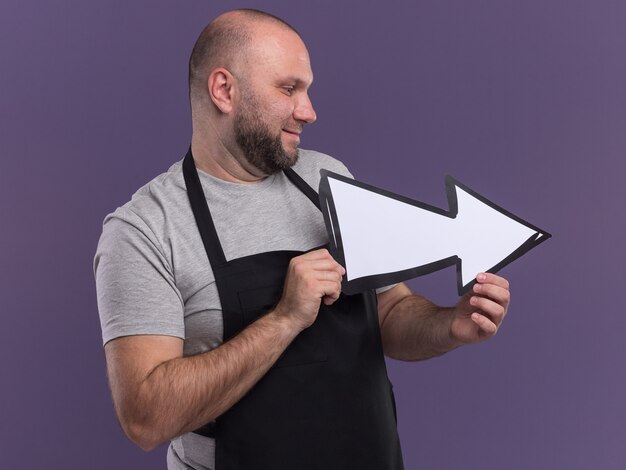 Sorridente guardando il barbiere maschio di mezza età laterale in uniforme che tiene il segno di direzione isolato sul muro viola