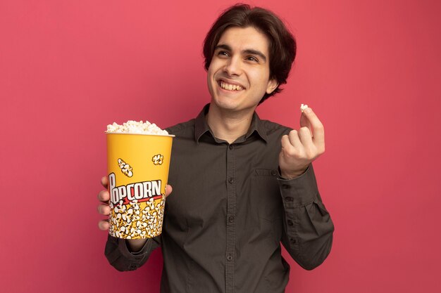 Sorridente guardando il lato giovane bel ragazzo che indossa una maglietta nera che tiene un secchio di popcorn con popcorn pace isolato sul muro rosa