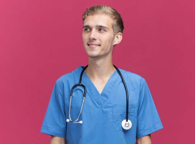 Sorridente guardando il lato giovane medico maschio che indossa l'uniforme del medico con lo stetoscopio isolato sulla parete rosa con spazio di copia