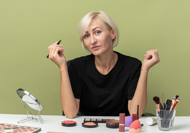 Sorridente guardando la telecamera la giovane bella ragazza si siede al tavolo con gli strumenti per il trucco tenendo l'eyeliner isolato su sfondo verde oliva