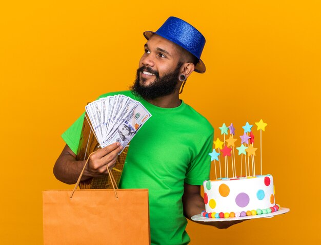 Sorridente guardando lato giovane ragazzo afro-americano che indossa un cappello da festa con regali e torta con contanti