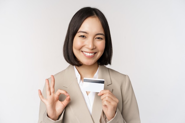 Sorridente impiegato d'ufficio asiatico donna aziendale che mostra carta di credito e segno giusto raccomandando banca in piedi su sfondo bianco in abito beige