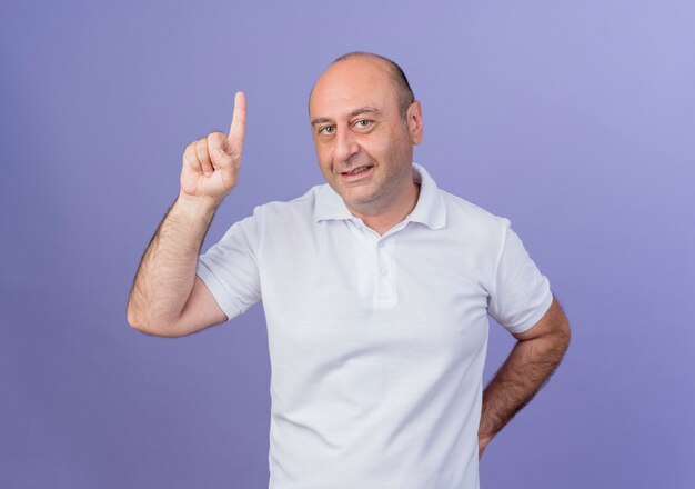 Sorridente imprenditore maturo casual mantenendo la mano dietro la schiena e alzando il dito isolato su sfondo viola