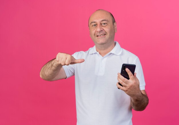 Sorridente imprenditore maturo casual tenendo e indicando il telefono cellulare isolato su sfondo rosa con copia spazio