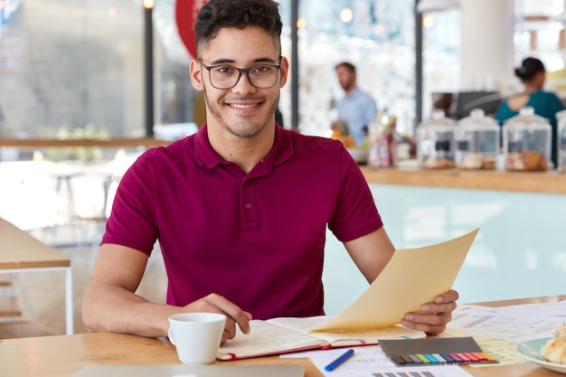 Sorridente imprenditore soddisfatto dei dati tiene documenti, indossa abiti casual, si prepara per il seminario di formazione, legge le informazioni necessarie, analizza la documentazione, posa contro l'interno del caffè. Condizioni di lavoro