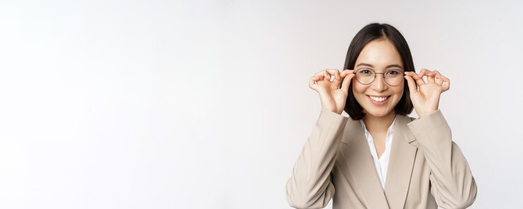 Foto gratuito sorridente imprenditrice asiatica cercando nuovi occhiali che indossano occhiali in piedi in tuta su sfondo bianco