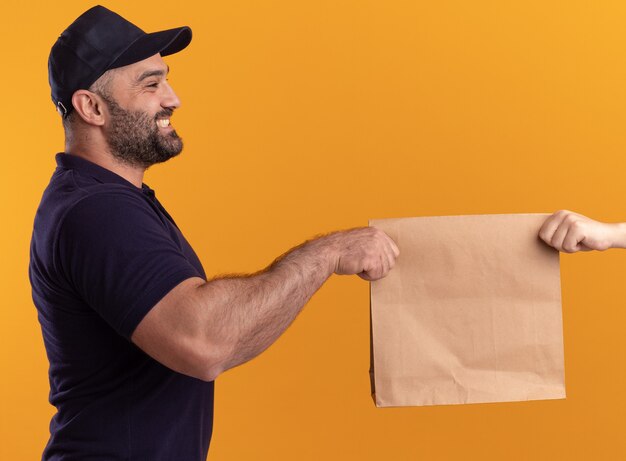 Sorridente in piedi nella vista di profilo fattorino di mezza età in uniforme e berretto che dà un pacchetto di cibo di carta al cliente isolato sul muro giallo