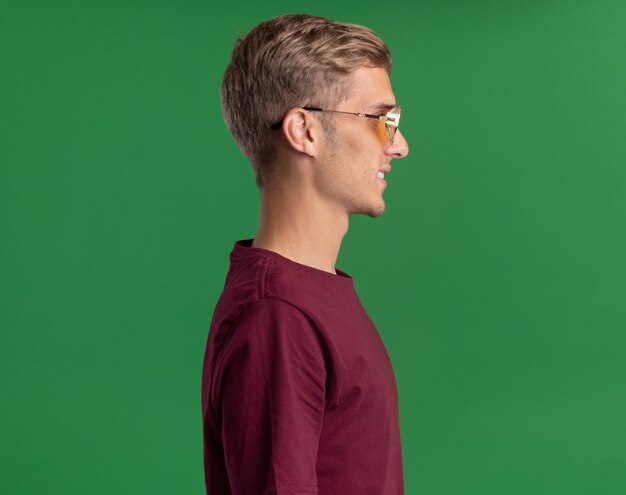 Sorridente in piedi nella vista di profilo giovane bel ragazzo che indossa una camicia rossa e occhiali isolati sul muro verde con spazio copia