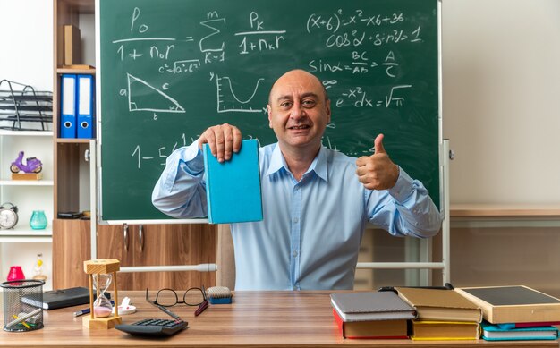 sorridente insegnante maschio di mezza età si siede al tavolo con materiale scolastico in possesso di libro che mostra il pollice in su in classe