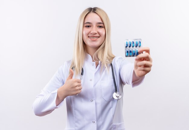 Sorridente medico giovane ragazza bionda che indossa stetoscopio e camice medico in parentesi graffa dentale azienda pillole il suo pollice in alto su sfondo bianco isolato