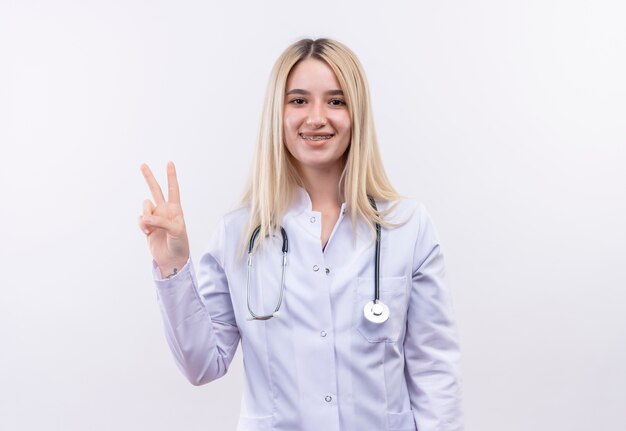 Sorridente medico giovane ragazza bionda che indossa uno stetoscopio e camice medico in parentesi graffa dentale che mostra gesto di pace isolato su sfondo bianco