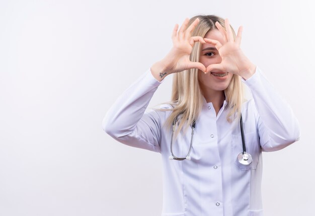 Sorridente medico giovane ragazza bionda che indossa uno stetoscopio e camice medico in parentesi graffa dentale che mostra il gesto del cuore su sfondo bianco isolato