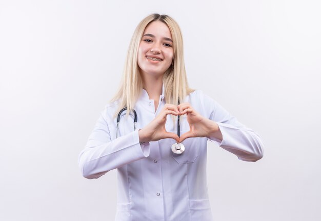 Sorridente medico giovane ragazza bionda che indossa uno stetoscopio e camice medico in parentesi graffa dentale che mostra il gesto del cuore su sfondo bianco isolato