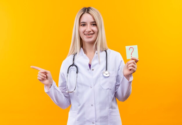 Sorridente medico giovane ragazza che indossa uno stetoscopio in abito medico e tutore dentale tenendo la carta punto interrogativo punti a lato su sfondo giallo isolato