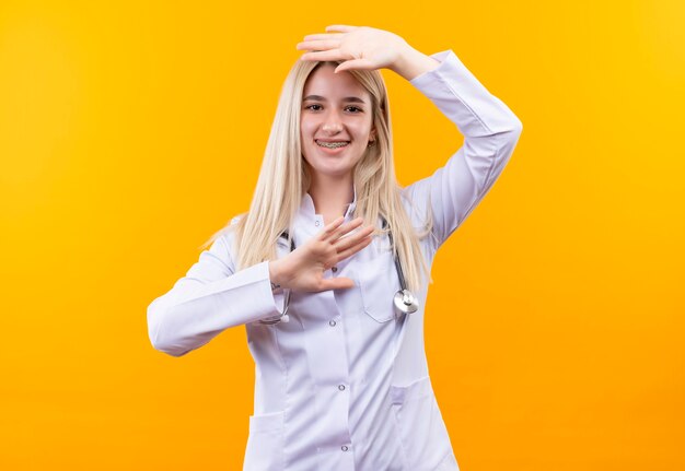 Sorridente medico giovane ragazza indossa stetoscopio in abito medico e tutore dentale mise la mano sulla testa altro sotto la testa su sfondo giallo isolato