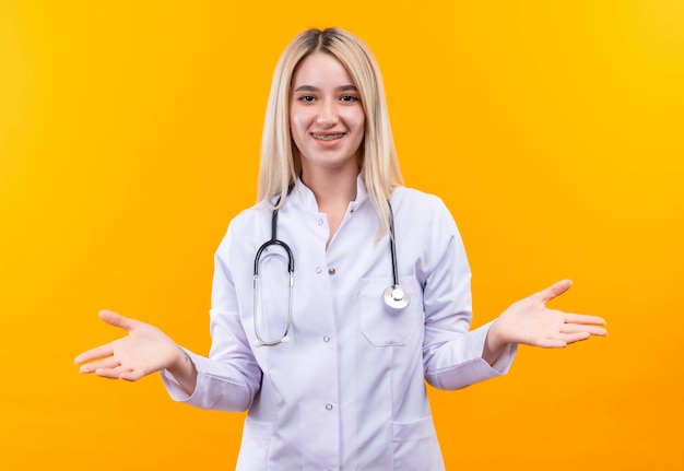 Sorridente medico giovane ragazza indossa uno stetoscopio in camice medico e parentesi graffe dentale diffonde le mani su sfondo giallo isolato