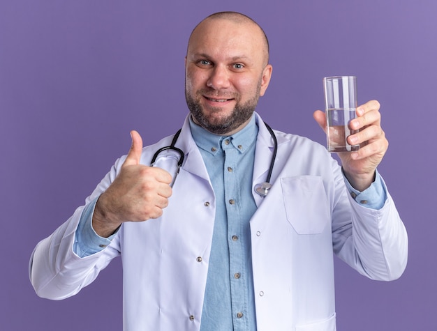 Sorridente medico maschio di mezza età che indossa una tunica medica e uno stetoscopio che tiene in mano un bicchiere d'acqua che mostra il pollice in su isolato sul muro viola