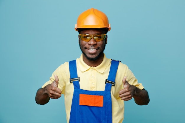 Sorridente mostrando pollice in alto giovane costruttore afroamericano in uniforme isolato su sfondo blu