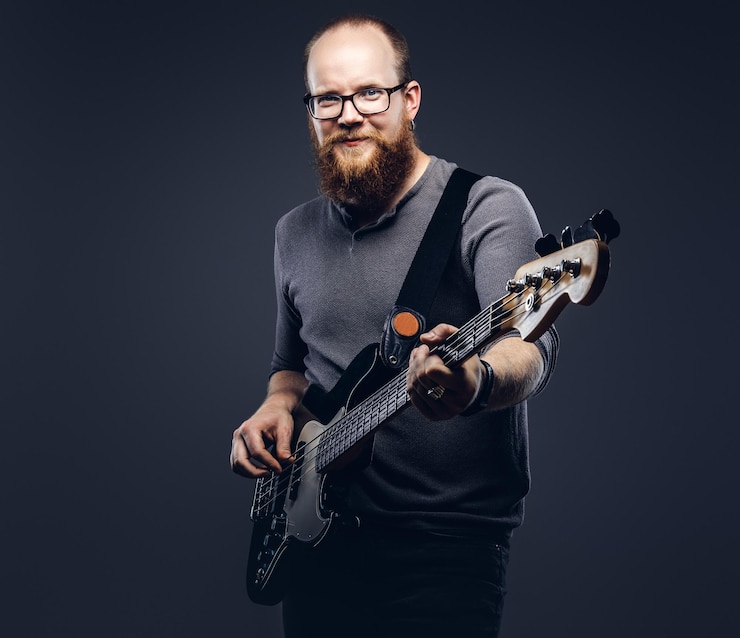 Foto gratuito sorridente musicista maschio con la barba rossa che indossa occhiali vestito con una t-shirt grigia che suona sulla chitarra elettrica. isolato su sfondo grigio.
