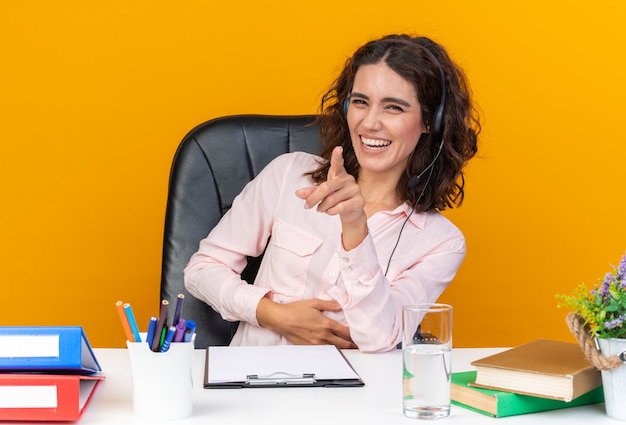 Sorridente operatore di call center femminile caucasica sorridente sulle cuffie seduto alla scrivania con strumenti da ufficio che puntano davanti