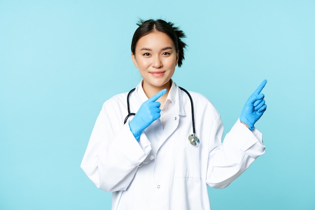 Sorridente operatore sanitario asiatico, indossando guanti sterili e uniforme, indicando a destra, dimostrando grafico, pubblicità ospedaliera, sfondo blu