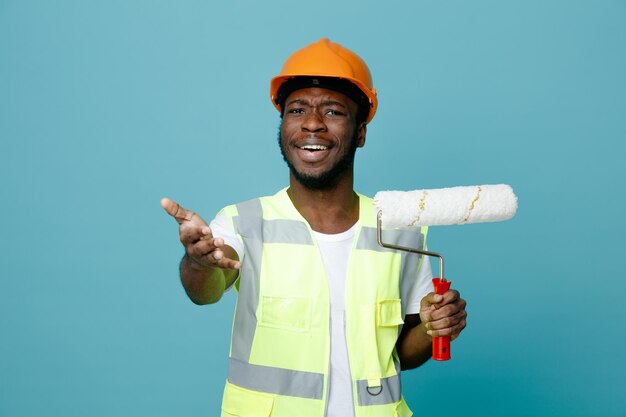Sorridente porgendo la mano alla telecamera giovane costruttore afroamericano in uniforme tenendo la spazzola a rullo isolata su sfondo blu