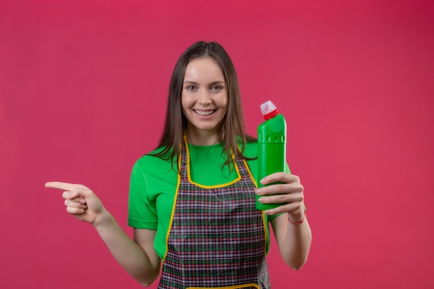 Sorridente pulizia ragazza giovane indossa uniforme azienda detergente punta il dito a lato su sfondo rosa isolato