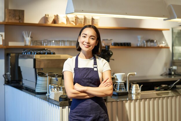 Sorridente ragazza asiatica barista in piedi vicino al bancone nella caffetteria che indossa il grembiule aziendale incrocia le mani e guarda