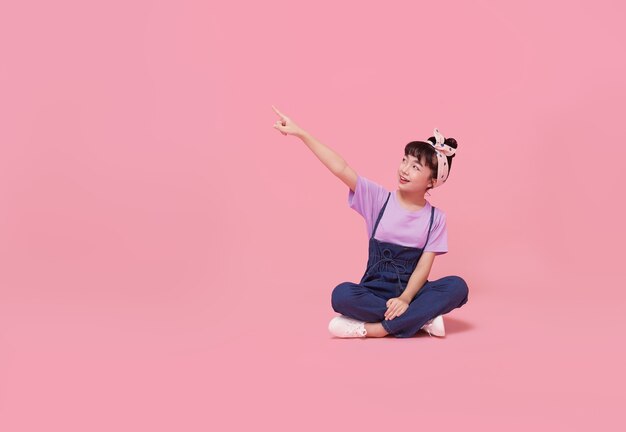 Sorridente ragazza asiatica del bambino che punta il dito su uno spazio vuoto accanto a una parete isolata rosa.