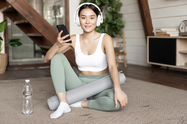 Sorridente ragazza asiatica in cuffie che indossa abbigliamento sportivo ascoltando musica tenendo il telefono cellulare e lavora...