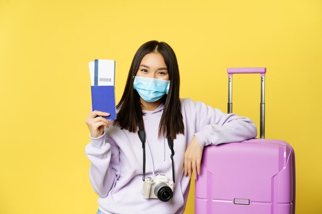Sorridente ragazza asiatica, turista in maschera medica, in posa vicino alla valigia, mostrando il passaporto, i biglietti per il viaggio di vacanza, in piedi su sfondo giallo