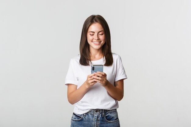 Sorridente ragazza attraente utilizzando il telefono cellulare e guardando lo schermo soddisfatto.