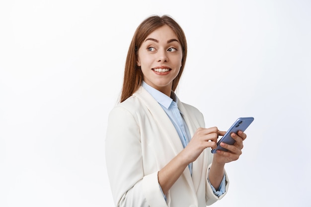 Sorridente ragazza aziendale in tuta che utilizza il telefono cellulare, gira la testa dietro e guarda il testo promozionale, leggendo lo spazio della copia sul muro bianco, tenendo in mano lo smartphone