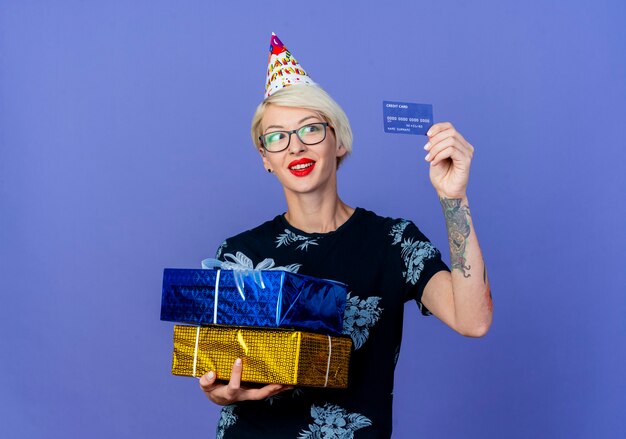 Sorridente ragazza bionda giovane partito con gli occhiali e berretto di compleanno che tiene scatole regalo e carta di credito guardando carta isolato su sfondo viola con spazio di copia